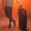 man in smoky, orange lit studio, holding a rolling luggage handle with edx backpack attached to it