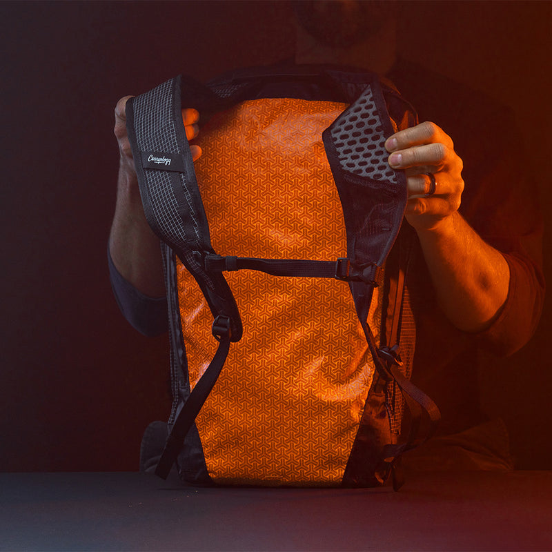 Man in dark studio, holding edx backpack straps, displaying back orange panel