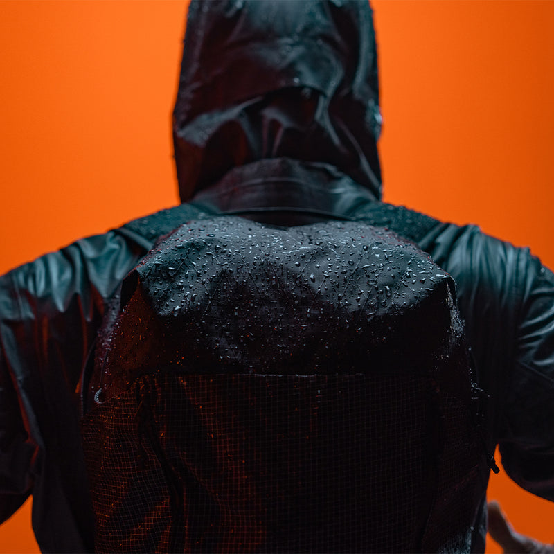 Person in raincoat on bright orange background, wearing black backpack covered in water droplets