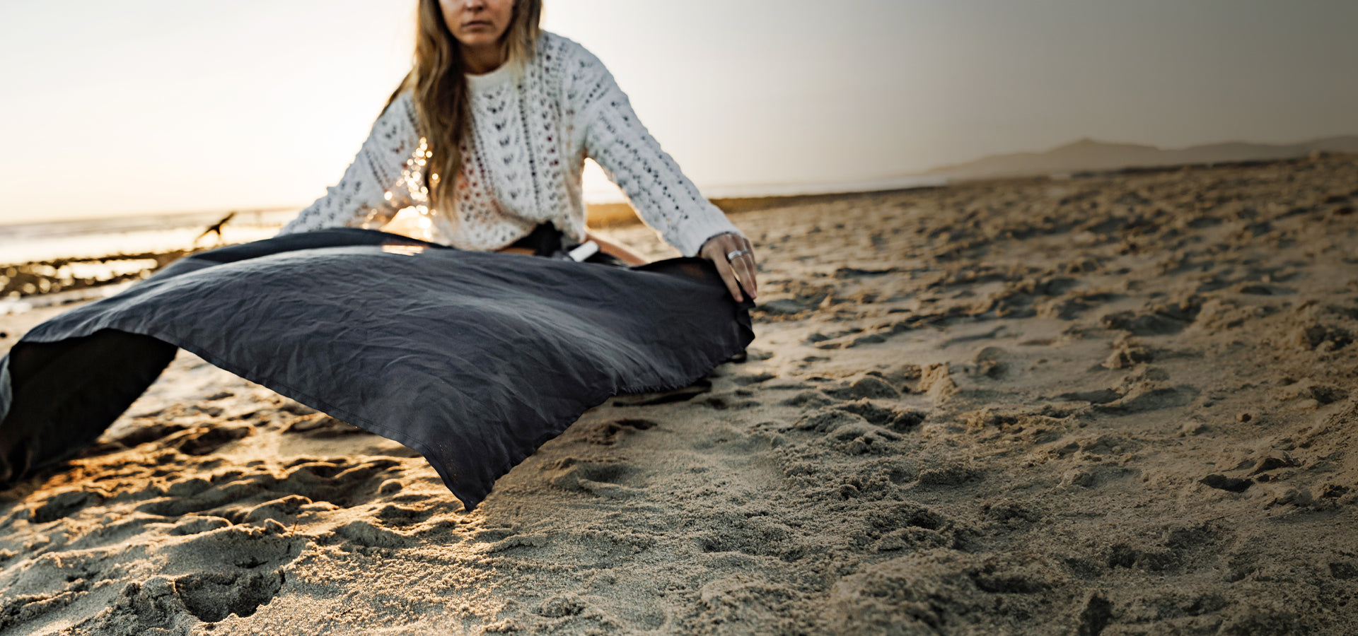 Woman in white sweater, laying out gray nanodry towel on sandy beach