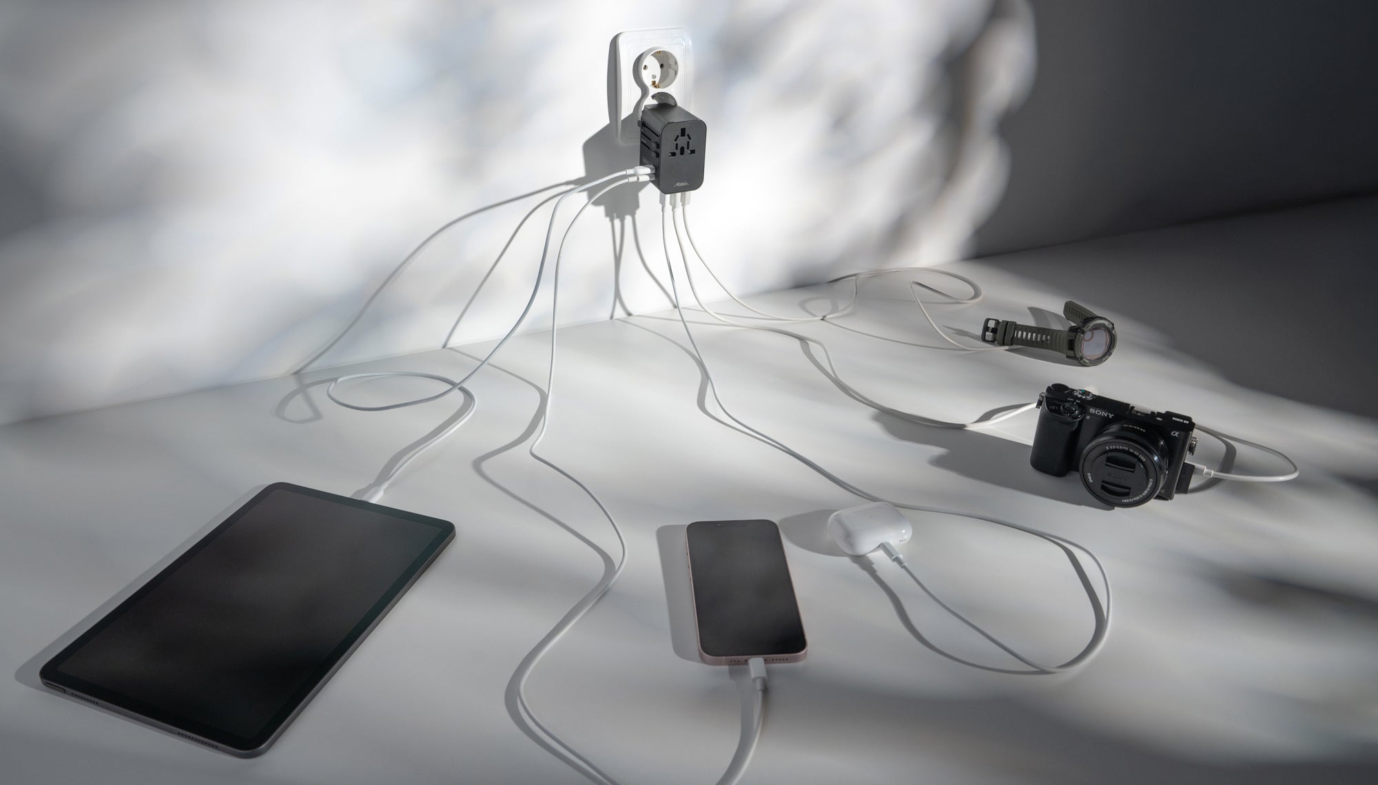 Global Travel Adapter plugged into EU outlet on white background. 5 devices are plugged into the adapter: ipad, phone, earbuds, camera, and smart watch