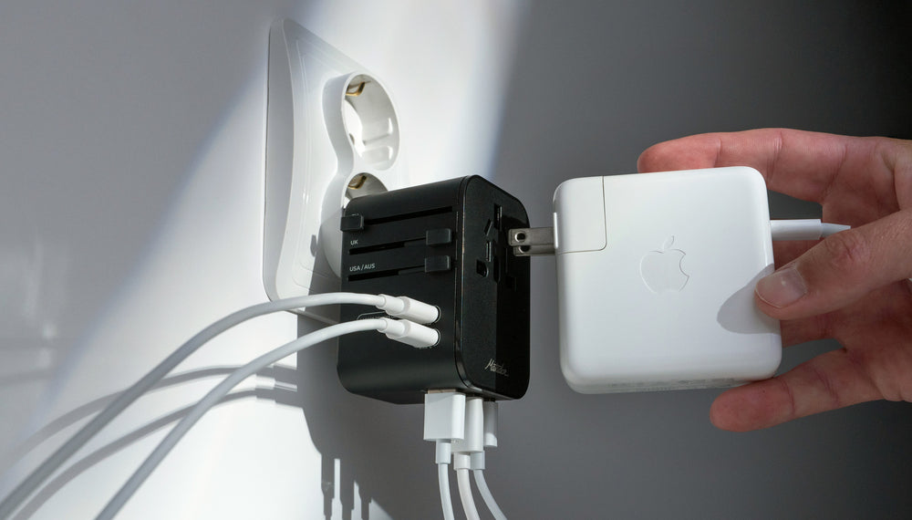 Close up view of Global Travel Adapter plugged into EU outlet on white wall with 5 white power cords plugged in. A hand is plugging in an apple laptop charger to the AC socket