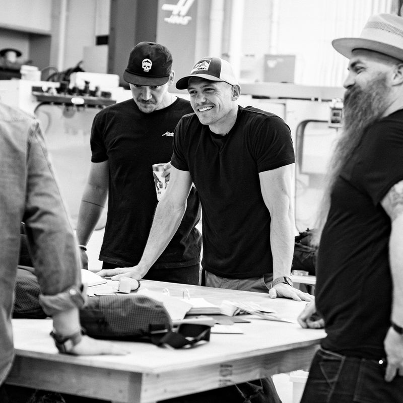 black and white photo: group of men smiling around a workbench covered in fabric and swatch samples
