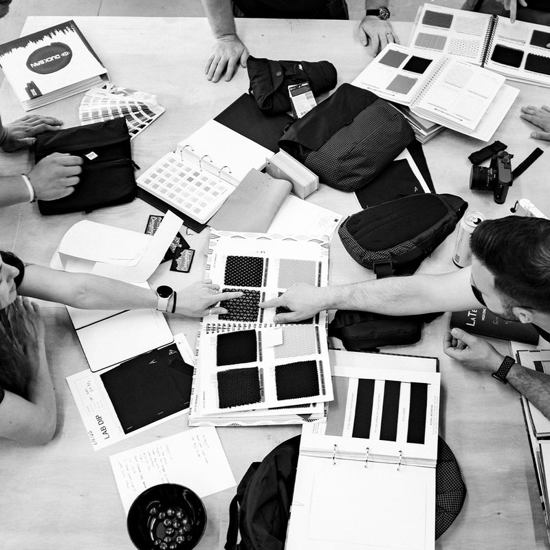 black and white image of people pointing at fabric swatches on a messy table of design supplies
