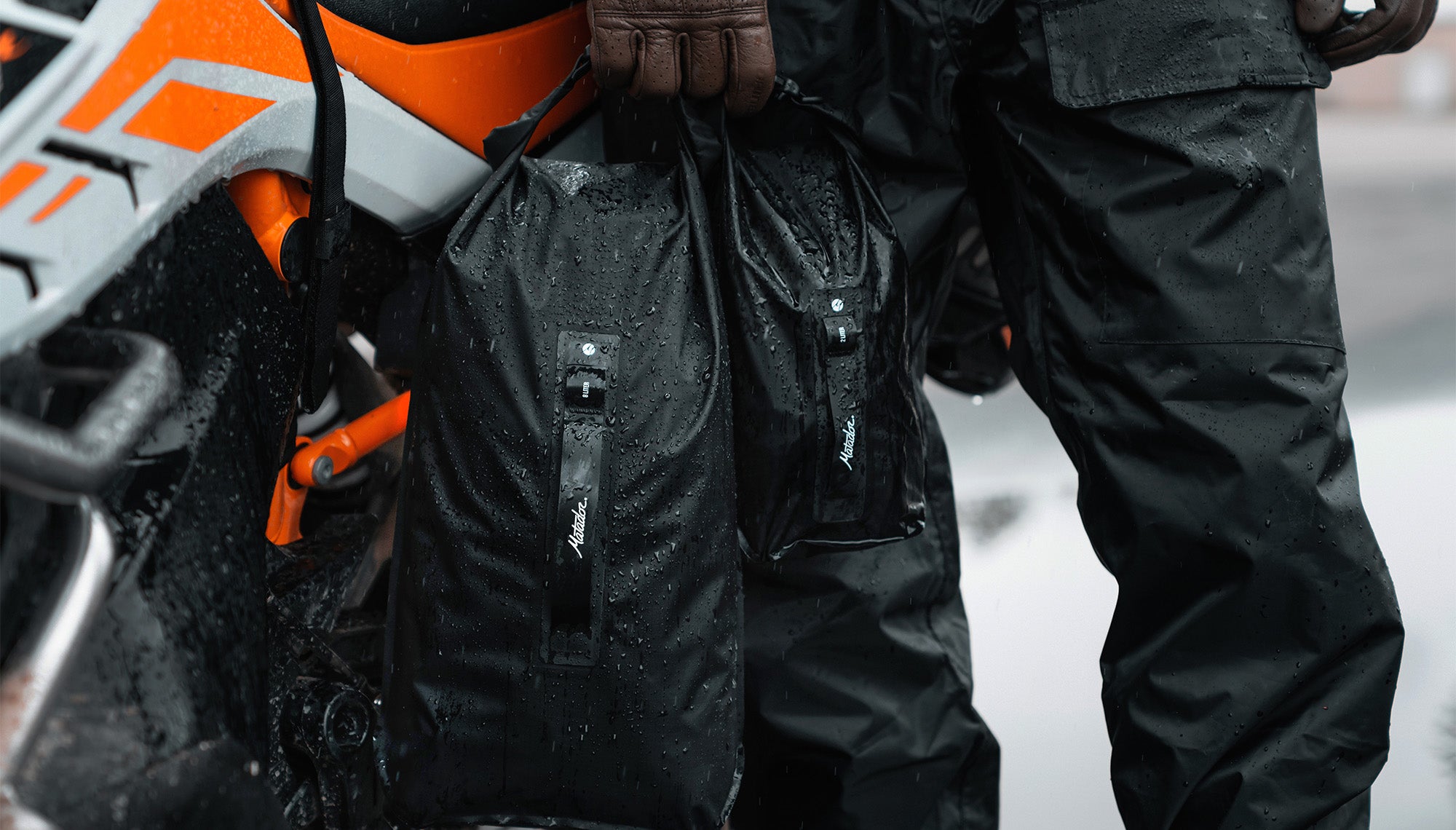 Person standing next to motorcycle, holding wet 8L and 2L dry bags 