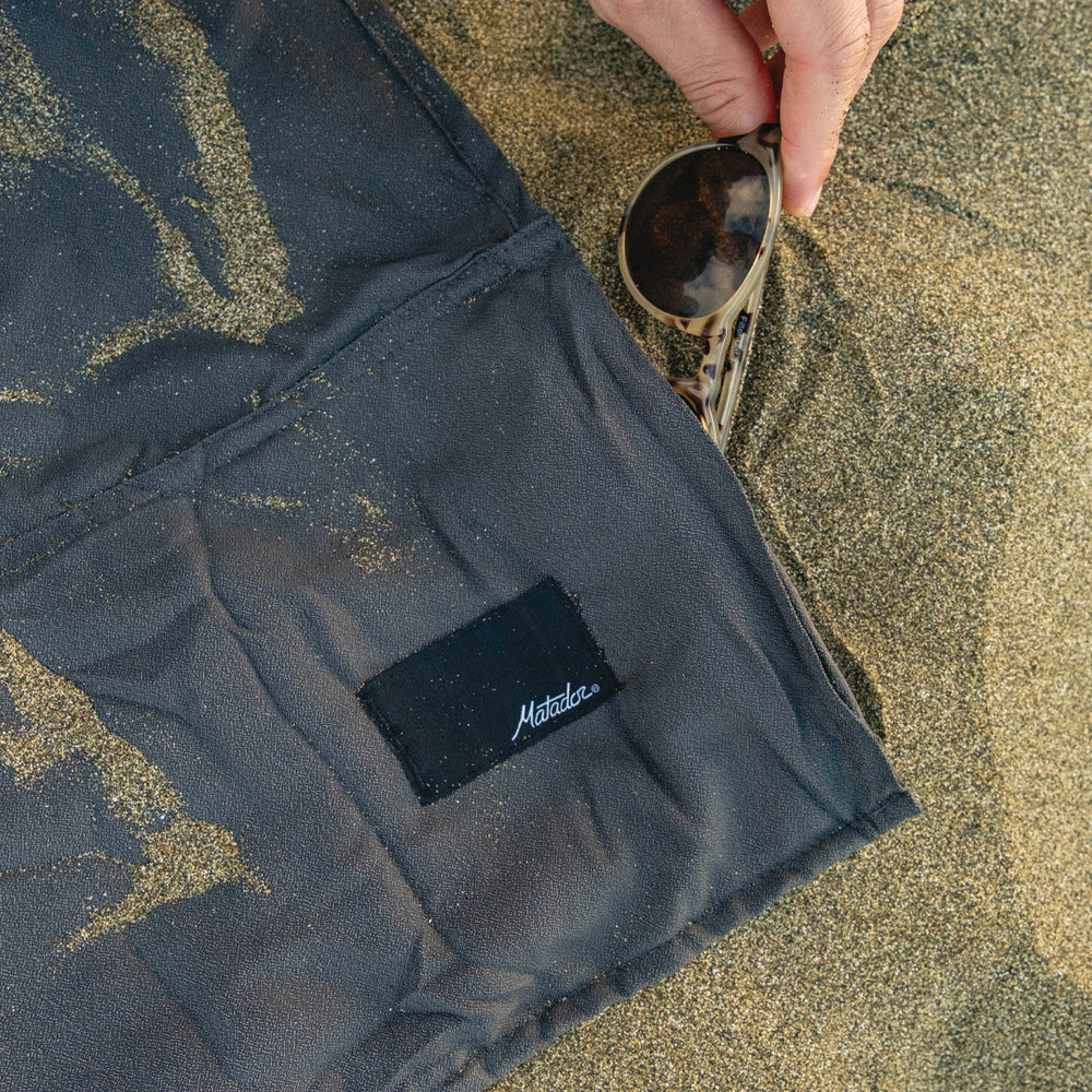 close up shot of nanodry beach towel in the sand with hand placing sunglasses in the pocket