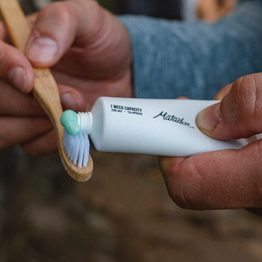 hand squeezing toothpaste out of white toothpaste tube onto bamboo toothbrush